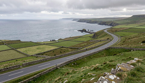 An elevated view over the Isle of Man from the mountain road, green fields and dry stone walls falling away to the sea under a grey Manx sky.