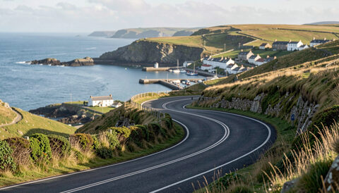 A tight hairpin bend on a hillside road above a harbour town, dark tarmac curving sharply, moorland and the sea beyond.