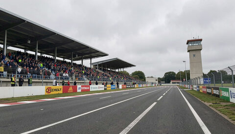 The TT Grandstand and start line on Glencrutchery Road with a packed grandstand, banners and a timing tower.